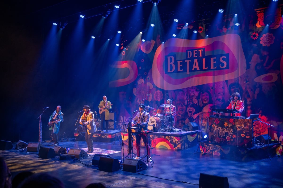 Det Betales live på scenen i blått lys med hele bandet og Beatles-bakteppet